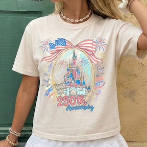K&ouml;nnte beinhalten: Cremefarbenes T-Shirt mit einem farbenfrohen Aufdruck eines Schlosses in einem ovalen Rahmen, mit einer Schleife aus der amerikanischen Flagge. Feuerwerk und der Text "250th Anniversary" sind ebenfalls abgebildet. Das Shirt ist ein Freizeitartikel.