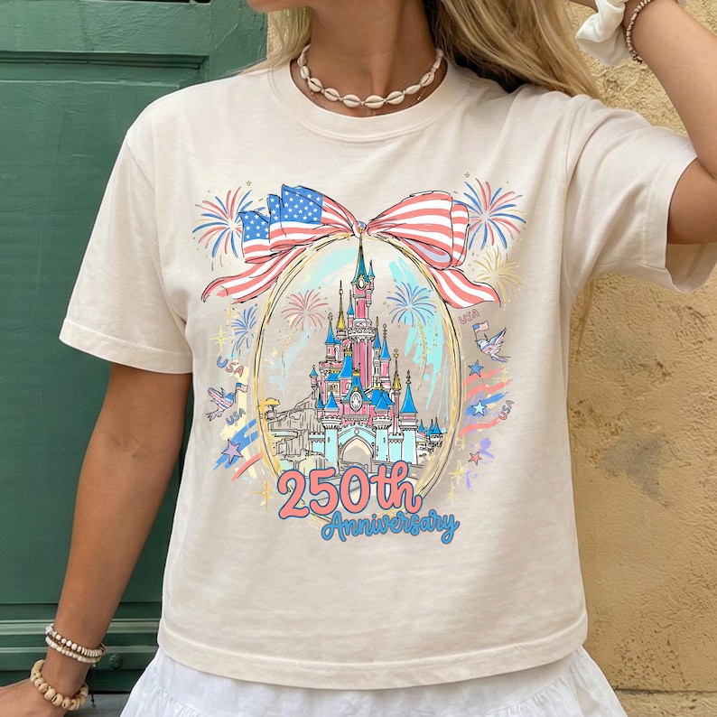 K&ouml;nnte beinhalten: Cremefarbenes T-Shirt mit einem farbenfrohen Aufdruck eines Schlosses in einem ovalen Rahmen, mit einer Schleife aus der amerikanischen Flagge. Feuerwerk und der Text "250th Anniversary" sind ebenfalls abgebildet. Das Shirt ist ein Freizeitartikel.