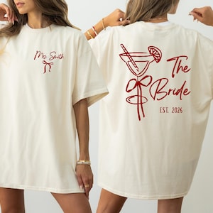 Peut inclure: T-shirt crème avec texte et graphiques rouges. Le devant affiche "Mrs. Smith" et un nœud. Le dos présente un verre à cocktail, les mots "The Bride" et "EST. 2026." Coupe ample et décontractée.