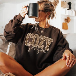 May include: A dark brown sweatshirt with the words "COZY season" in a leopard print design. The person in the image is holding a black mug. The sweatshirt has long sleeves and a crew neck.