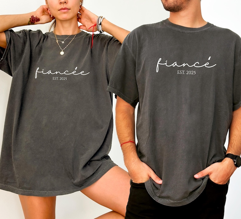 May include: Two charcoal gray t-shirts with the word "fianc&eacute;e" in cursive script and the text "EST. 2025". The shirts are a relaxed fit and appear to be made of a soft material. The shirts are worn by two people.