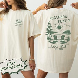 May include: An off-white t-shirt featuring a small green graphic on the front left chest and a large green graphic on the back. The back design depicts two chairs by a lake, trees, a sun, and the text: ANDERSON FAMILY LAKE TRIP EST. 2026.