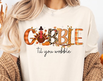 May include: Cream-colored t-shirt with a Thanksgiving-themed graphic. The design features the word "GOBBLE" in orange plaid, leopard print, and brown lettering, with a turkey and pumpkins. The phrase "Til you wobble" is written below.