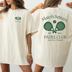 May include: An off-white t-shirt featuring a small green graphic on the front chest and a large green graphic on the back. The back design displays two crossed padel rackets, a ball, and the text Match Setters PADEL CLUB AUSTIN, TEXAS EST. 2023.