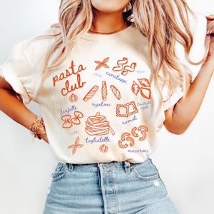 Pu&ograve; includere: T-shirt color crema con un'illustrazione di pasta rossa e blu. Le parole "pasta club" sono in corsivo rosso. Varie forme di pasta sono etichettate, tra cui farfalle, rigatoni e ravioli. La maglietta &egrave; abbinata a jeans blu.