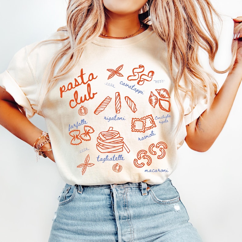Pu&ograve; includere: T-shirt color crema con un'illustrazione di pasta rossa e blu. Le parole "pasta club" sono in corsivo rosso. Varie forme di pasta sono etichettate, tra cui farfalle, rigatoni e ravioli. La maglietta &egrave; abbinata a jeans blu.