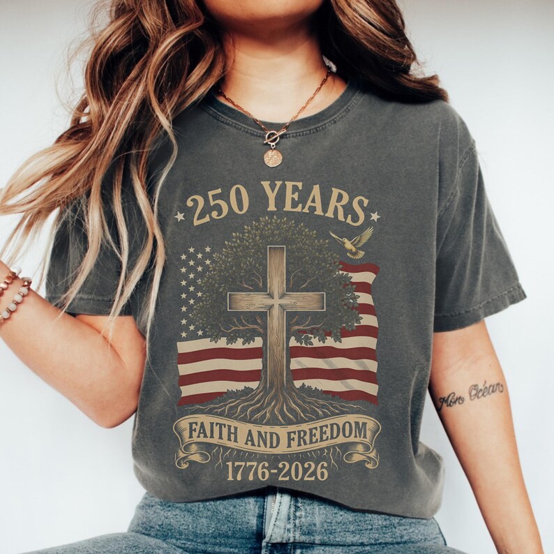 May include: Dark gray t-shirt with a graphic design celebrating 250 years. The design includes an American flag, a cross, a tree, and the text "Faith and Freedom 1776-2026". A flying bird is also featured in the design.