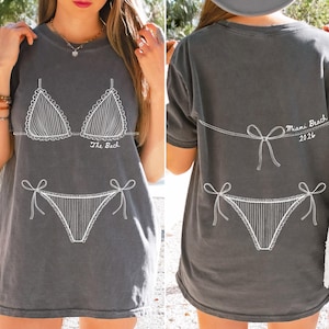 May include: Dark gray t-shirt featuring a white line drawing of a bikini. The front of the shirt says "The Back". The back of the shirt has the text "Miami Beach 2026".