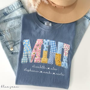 Puede incluir: Una camiseta azul Comfort Colors con la palabra "MINI" en letras de patchwork. Las letras están decoradas con estampados florales y de cuadros. Debajo de la palabra hay nombres. Un sombrero beige y unos vaqueros azules están en el fondo.