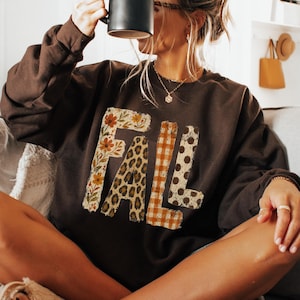 May include: A dark brown long-sleeve crewneck sweatshirt featuring the word FALL printed across the chest. Each letter has a distinct pattern: the F has a floral print, the A has a leopard print, the L has an orange plaid, and the L has brown polka dots.