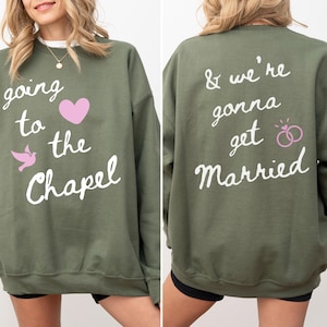 May include: Olive green sweatshirt with the text "going to the Chapel" and a pink heart and dove graphic on the front. The back of the sweatshirt says "& we're gonna get Married" with a pink rings graphic. A casual wedding-themed top.