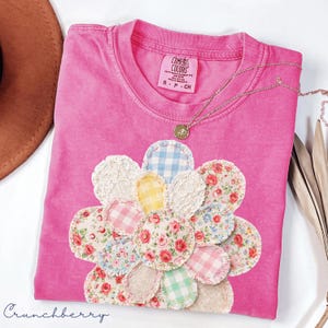 Puede incluir: Una camiseta rosa Comfort Colors con un diseño floral de aplique. El aplique presenta varios círculos de tela estampada, incluyendo flores, cuadros y encaje. Un collar de oro con un colgante descansa sobre la camisa. La palabra "Crunchberry" está escrita en cursiva en la parte inferior.