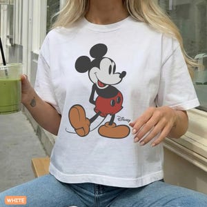 May include: White t-shirt featuring a classic Mickey Mouse graphic. The cartoon character is depicted in his iconic pose, wearing red shorts and brown shoes. The Disney logo is visible. The word "WHITE" is at the bottom.