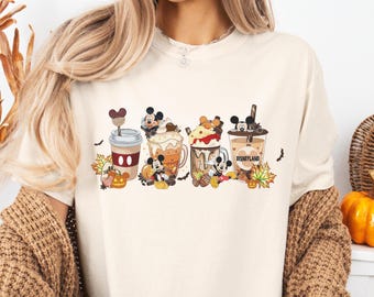 Può includere: T-shirt color crema con una grafica colorata di bevande autunnali a tema Mickey Mouse. Il design include tazze di caffè e zucche, con la scritta "DISNEYLAND" visibile su una tazza. La maglietta ha scollo tondo e maniche corte.