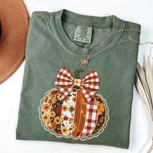 Puede incluir: Una camiseta verde salvia Comfort Colors con un diseño decorativo de calabaza. La calabaza está compuesta por varios patrones, incluyendo florales y cuadros, con un gran lazo a cuadros. Un collar de oro con un colgante está drapeado sobre la camisa.