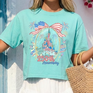 K&ouml;nnte beinhalten: Hellblaues T-Shirt mit einem farbenfrohen Schloss-Motiv in einem ovalen Rahmen, Feuerwerk und einer Schleife mit amerikanischer Flagge. Der Text "250th Anniversary" steht darunter. Das Shirt ist im Cropped-Stil.