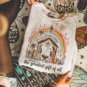 May include: Beige sweatshirt featuring a colorful illustration of the Nativity scene with a rainbow, stars, and palm trees. The text "Matthew 1:21" and "the greatest gift of all" are printed below the image. The sweatshirt is held by a person.