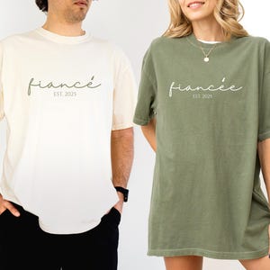 May include: Two oversized t-shirts, one cream and one olive green. Each shirt has the word "fianc&eacute;" in a script font, with "EST. 2025" below it. The shirts are likely for a couple, possibly for an engagement announcement or wedding.