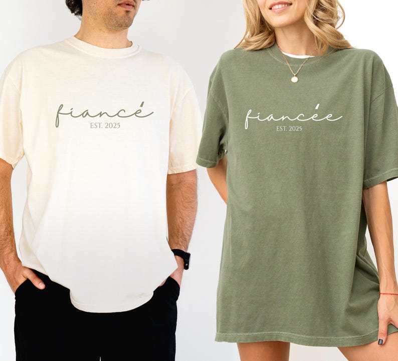 May include: Two oversized t-shirts, one cream and one olive green. Each shirt has the word "fianc&eacute;" in a script font, with "EST. 2025" below it. The shirts are likely for a couple, possibly for an engagement announcement or wedding.