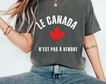 May include: Dark gray t-shirt with the text "LE CANADA" above a red maple leaf and the text "N'EST PAS À VENDRE" below. The shirt is a casual style.