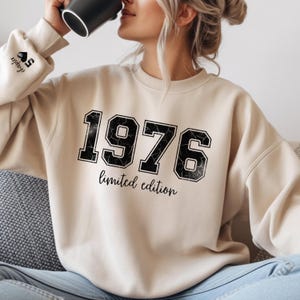 Può includere: Felpa beige con l'anno "1976" in nero, bordato di bianco, e le parole "limited edition" sotto. La manica sinistra ha il testo "chapter 50" e un simbolo di carta da gioco. Una persona tiene una tazza nera.