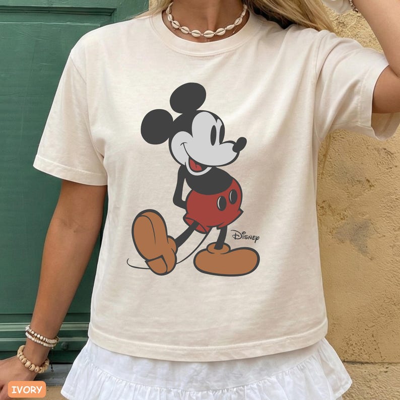 May include: Ivory-colored t-shirt featuring a classic Mickey Mouse graphic. The cartoon character is depicted in black, red, and brown, with the Disney logo printed below. The shirt has a crew neck and short sleeves.