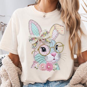Può includere: T-shirt color crema con un design colorato di coniglietto pasquale. Il coniglietto ha occhiali, un fiocco e dettagli floreali. Il design incorpora colori pastello e abbellimenti perlati, creando un aspetto stravagante e festoso.