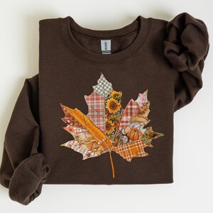 May include: A brown sweatshirt with a large maple leaf design. The leaf is made from fabric patches with autumn patterns, including sunflowers and plaid. The sweatshirt has a crew neck and long sleeves. The design evokes the season of autumn.