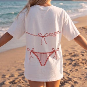 May include: White t-shirt with a red bikini design and the text "Palm Beach 2026". The graphic features a bikini top and bottom with bow details. The shirt is a casual, beach-themed apparel item.