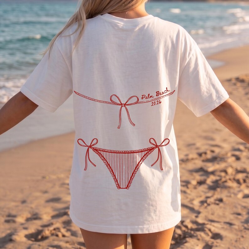 May include: White t-shirt with a red bikini design and the text "Palm Beach 2026". The graphic features a bikini top and bottom with bow details. The shirt is a casual, beach-themed apparel item.