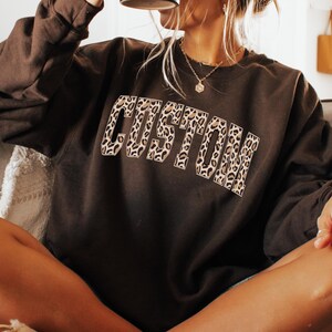 Op de afbeelding: Een donkerbruine sweatshirt met het woord "CUSTOM" in een luipaardprint ontwerp over de borst. De sweatshirt heeft lange mouwen en een ronde hals. De afbeelding is een close-up.
