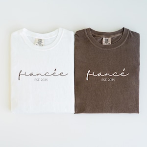 May include: Two folded t-shirts, one white and one brown, with the word "fianc&eacute;e" in a cursive font and the text "EST. 2025" printed below. The shirts are a comfortable, casual style.