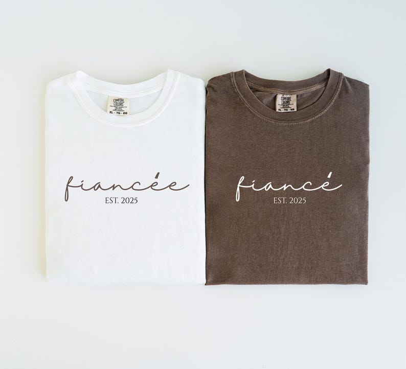 May include: Two folded t-shirts, one white and one brown, with the word "fianc&eacute;e" in a cursive font and the text "EST. 2025" printed below. The shirts are a comfortable, casual style.