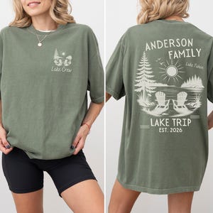 May include: An olive green short-sleeve t-shirt featuring a small graphic on the front left chest with two chairs, trees, and the text Lake Crew. The back displays a large graphic of two chairs by a lake, trees, sun, and text Anderson Family Lake Trip Est. 2026.