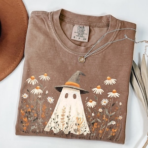 May include: Brown Comfort Colors t-shirt featuring a whimsical ghost wearing a witch hat, surrounded by daisies and foliage. A silver necklace with a pendant rests on the shirt. The shirt is size S-P-CH.