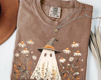 May include: Brown Comfort Colors t-shirt with a ghost in a witch hat design, set amongst daisies and plants. A silver necklace with a pendant is on the shirt. The shirt is size S-P-CH.