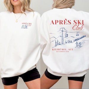 Könnte beinhalten: Ein weißes Sweatshirt mit Rundhalsausschnitt und einer kleinen rot-blauen Grafik auf der linken Brust, die "APRÈS SKI Club" mit Skiern zeigt. Die Rückseite zeigt eine große blaue Skilift-Illustration mit dem roten Text "APRÈS SKI Club KITZBÜHEL, AUT DECEMBER 2026".