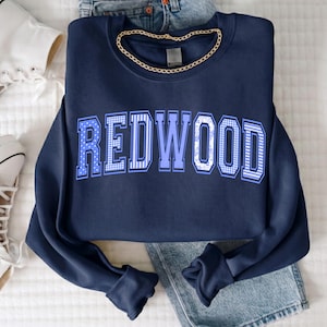 Puede incluir: Una sudadera azul marino de cuello redondo con la palabra REDWOOD en grandes letras de bloque azul claro. Cada letra tiene un patrón único, incluyendo lunares, rayas verticales y cuadros vichy. Una cadena dorada descansa en el cuello.