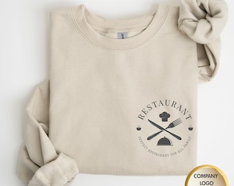 May include: Beige crewneck sweatshirt with a black restaurant logo. The logo features a chef's hat, crossed knife and fork, and the words "Restaurant" and "Perfect Restaurant for All Family". A matching scrunchie is next to the sweatshirt.