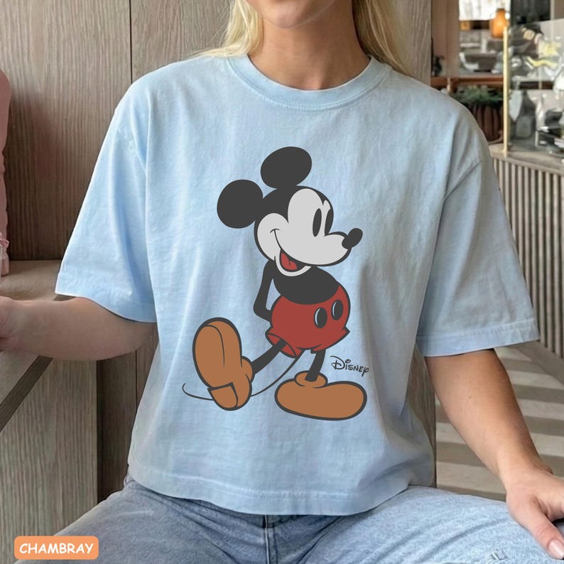 May include: Light blue t-shirt featuring a graphic of Mickey Mouse in a classic pose. The cartoon character is depicted in black, white, red, and brown. The Disney logo is printed below the character. The shirt is made of a soft material.