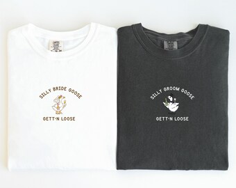 May include: Two crew neck t-shirts, one white and one charcoal grey. The white shirt displays a cartoon goose with a drink, and the text "SILLY BRIDE GOOSE GETT'N LOOSE". The grey shirt has a similar design with the text "SILLY GROOM GOOSE GETT'N LOOSE".