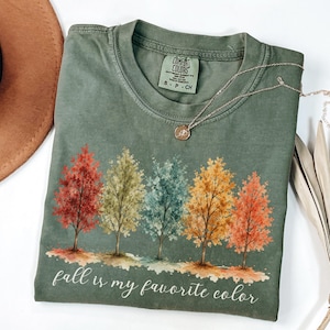 May include: An olive green short-sleeve t-shirt featuring a graphic of five watercolor-style trees in shades of red, green, blue, yellow, and orange. The text "fall is my favorite color" is printed in white script below the trees.