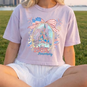K&ouml;nnte beinhalten: Helllila T-Shirt mit einem Schlossdesign in einem Oval, verziert mit einer Schleife aus der amerikanischen Flagge. Der Text "250th Anniversary" befindet sich unter dem Schloss. Feuerwerk und Sterne umgeben das Oval.