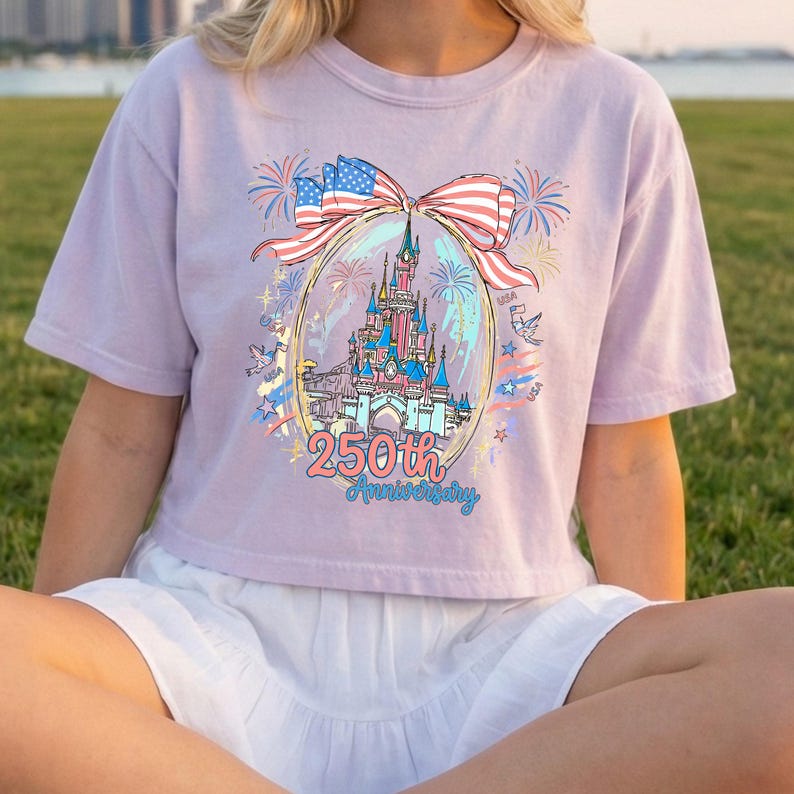 K&ouml;nnte beinhalten: Helllila T-Shirt mit einem Schlossdesign in einem Oval, verziert mit einer Schleife aus der amerikanischen Flagge. Der Text "250th Anniversary" befindet sich unter dem Schloss. Feuerwerk und Sterne umgeben das Oval.