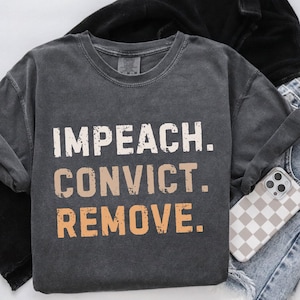 May include: Dark gray sweatshirt with the words "IMPEACH. CONVICT. REMOVE." in a distressed, layered font. The sweatshirt is folded, with a phone case featuring a checkered pattern visible in the corner.