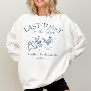 Op de afbeelding: Crèmekleurige sweatshirt met een blauwe print. De print toont de tekst "Last Toast On the Slopes", een martiniglas met een bruid, ski's en een boom. Daaronder staat "Emily's Bachelorette ASPEN, CO".