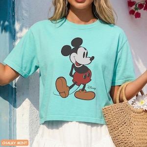May include: A light teal t-shirt featuring a graphic of Mickey Mouse. The cartoon character is depicted in his classic outfit: red shorts, brown shoes, and black ears. The Disney logo is printed below the character.