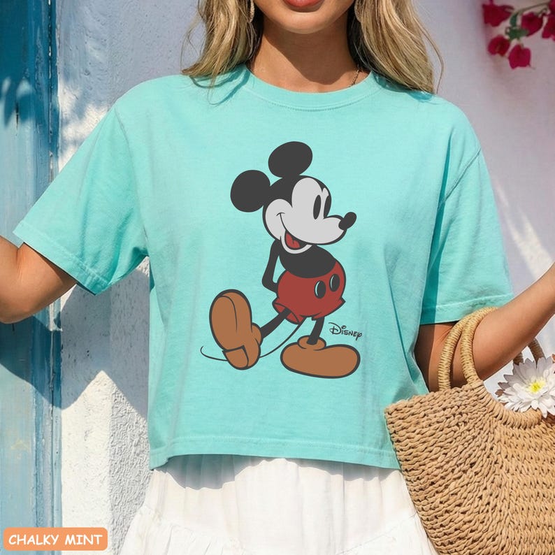 May include: A light teal t-shirt featuring a graphic of Mickey Mouse. The cartoon character is depicted in his classic outfit: red shorts, brown shoes, and black ears. The Disney logo is printed below the character.