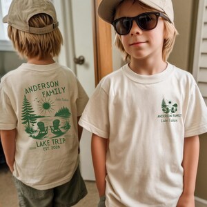 May include: A cream-coloured t-shirt featuring a green graphic of a lake scene with two chairs, pine trees, and the text Anderson Family Lake Trip Est. 2026 on the back, and a smaller matching graphic on the front left chest area.