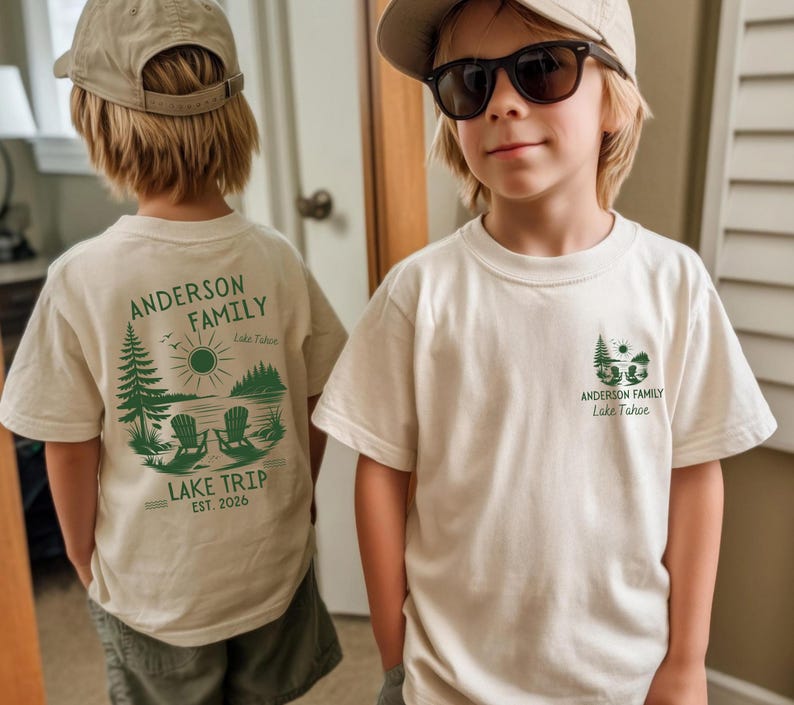 May include: A cream-coloured t-shirt featuring a green graphic of a lake scene with two chairs, pine trees, and the text Anderson Family Lake Trip Est. 2026 on the back, and a smaller matching graphic on the front left chest area.